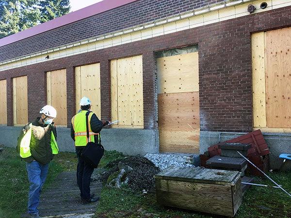 workers stand in front of a brick building that has holes for windows and a door, but they are filled with plywood