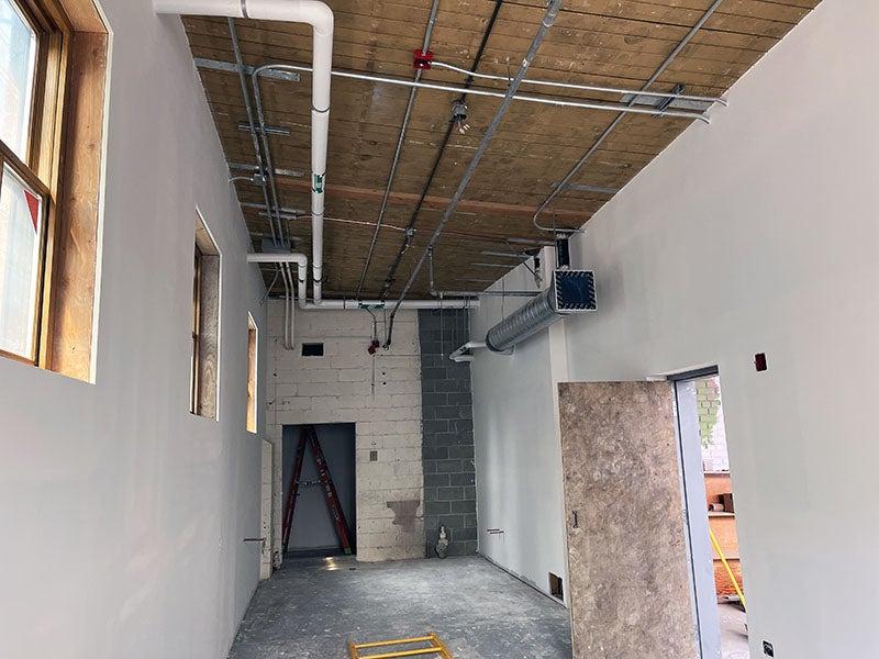 a room with piping at the ceiling, windows on the right side wall, and a door on the left side. the floor is concrete and the back wall is concrete masonry units